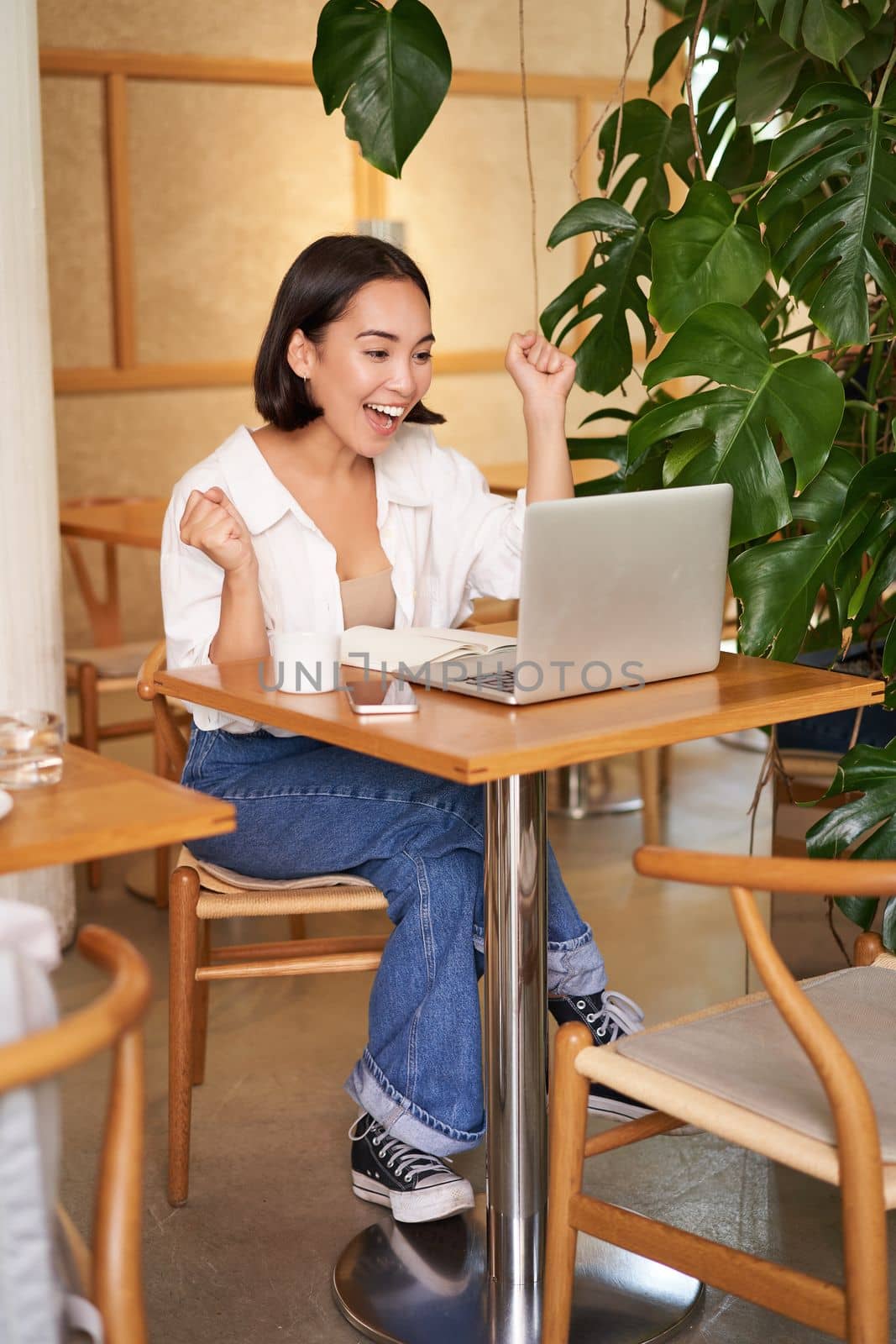 Enthusiastic asian girl celebrating, looking at laptop and triumphing, makes fist pumps with excited face, sits in cafe.