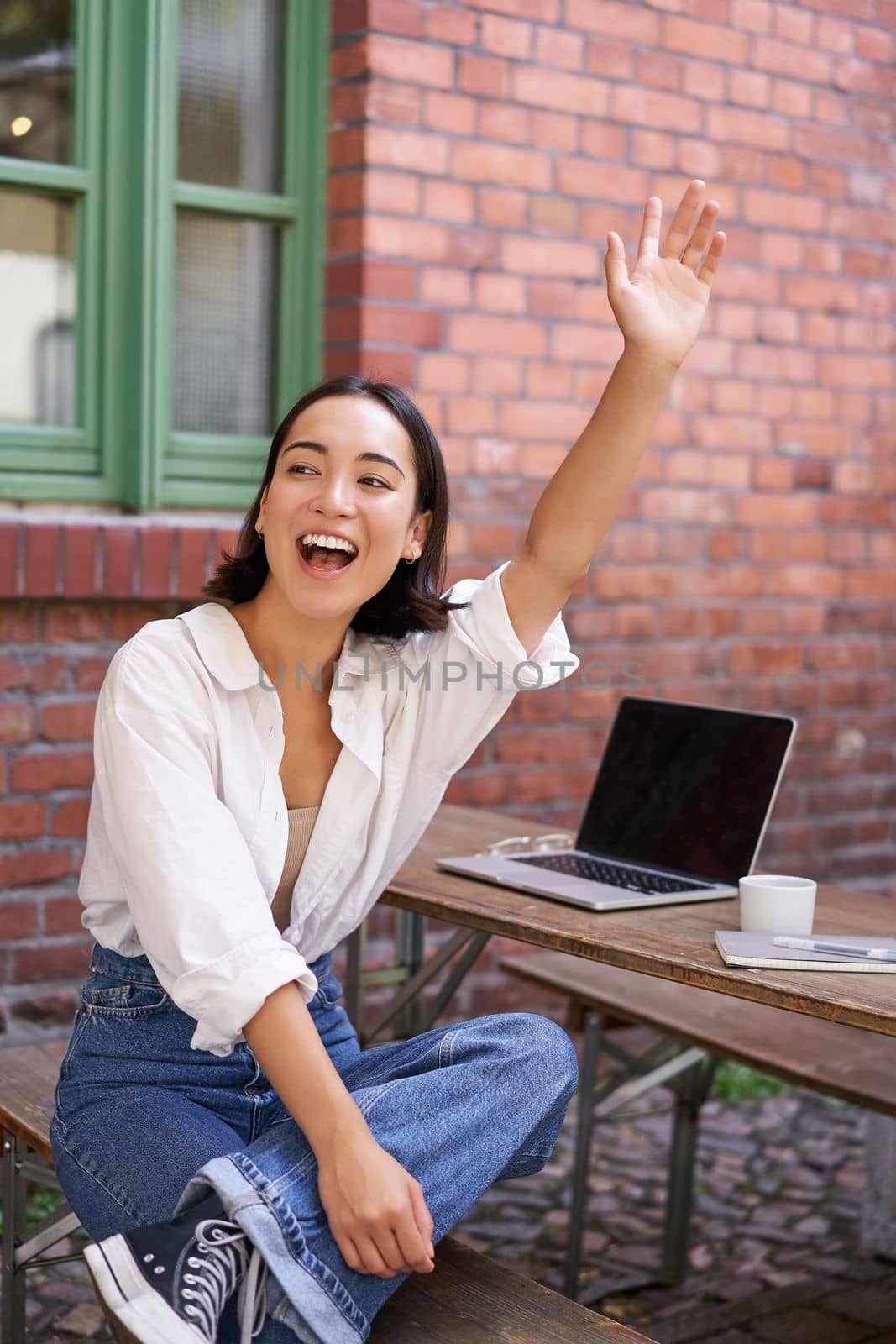 Vertical shot of happy girl attracting attention, waving hand at friend in cafe, sitting on bench with laptop.