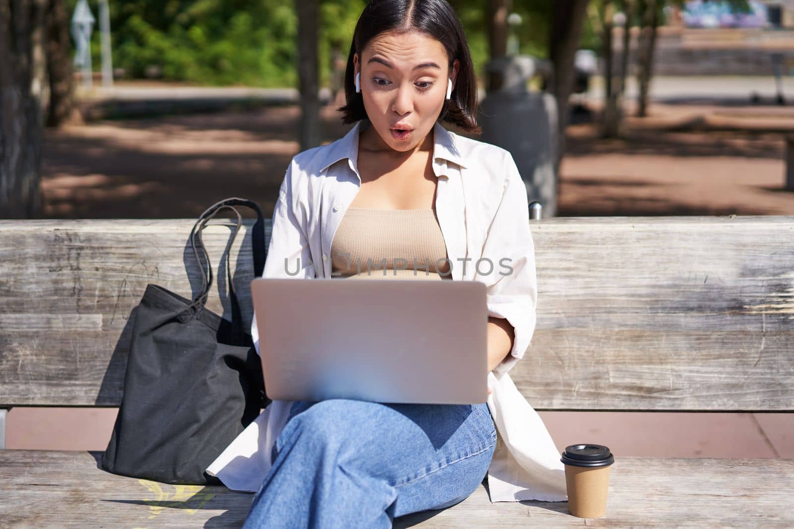 Happy asian girl chatting, having video call on laptop, sitting on bench in park and communicating, talking to someone online.