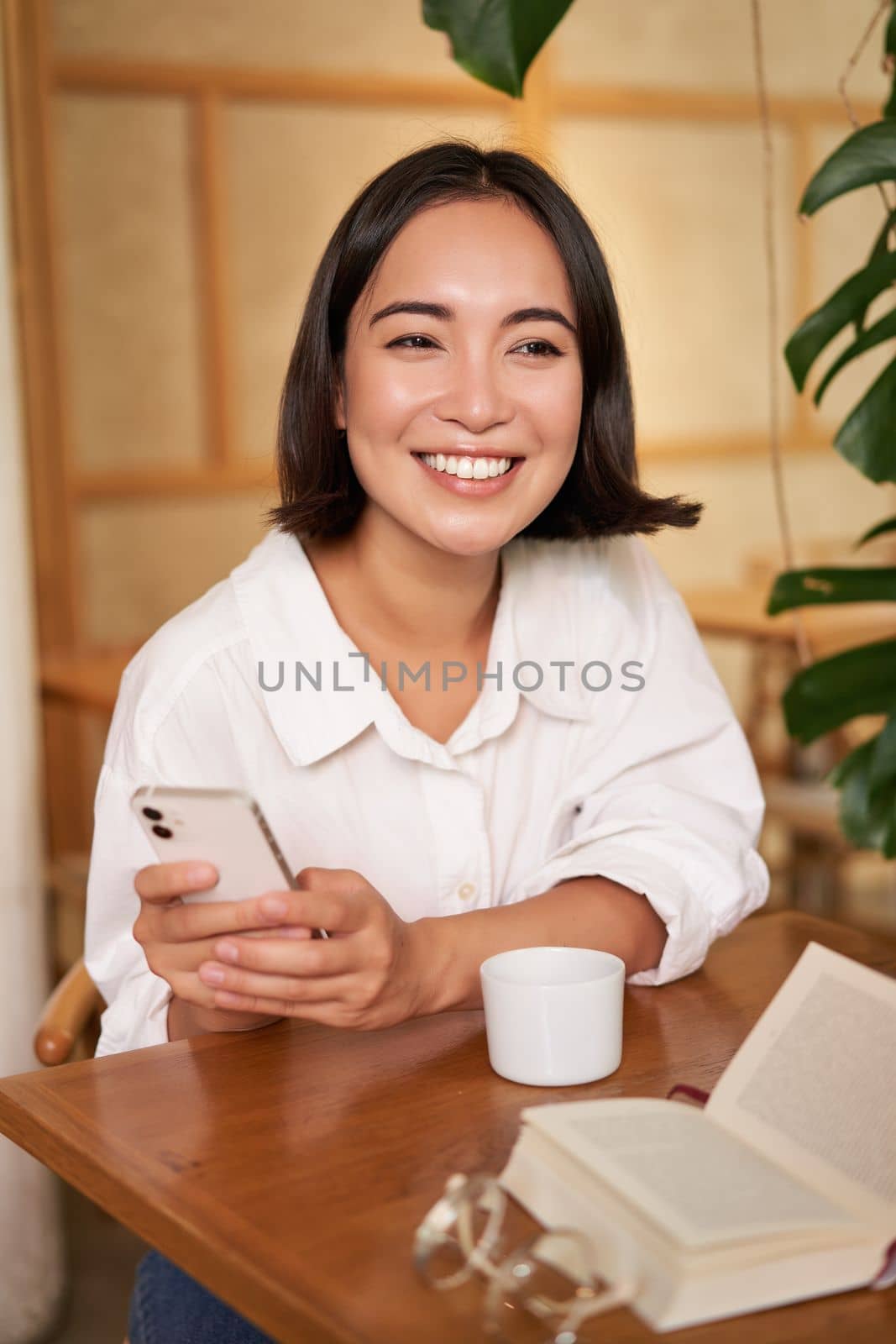 Portrait of modern girl sits with coffee in cafe, smiling while looking at smartphone, reading book in restaurant and using mobile phone app.