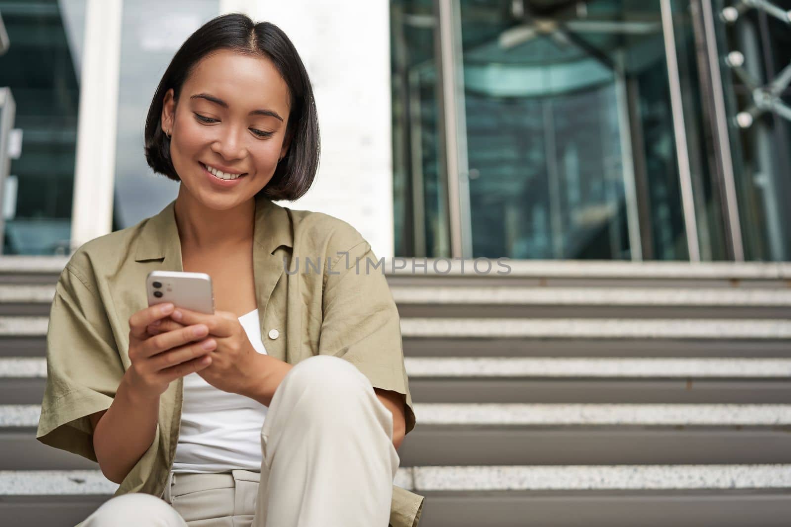 Smiling asian girl sits on stairs near building entrance, using mobile phone app. Happy young woman rests with smartphone in her hands outdoors by Benzoix