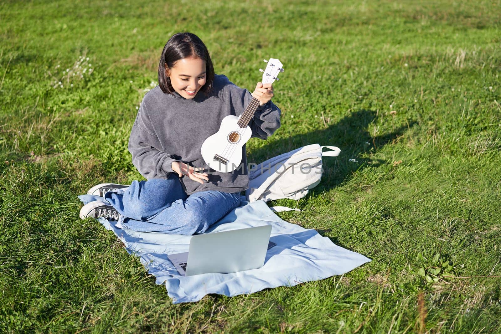 Cheerful asian girl, musician waves at her laptop, say hi to video chat, plays ukulele and records it on laptop, sits on grass in park by Benzoix