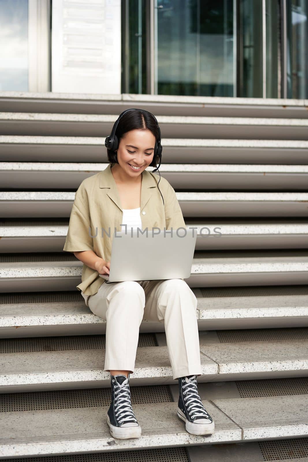 Smiling asian girl in headphones, works on laptop, digital nomad using computer on remote, sitting on stairs outdoors.