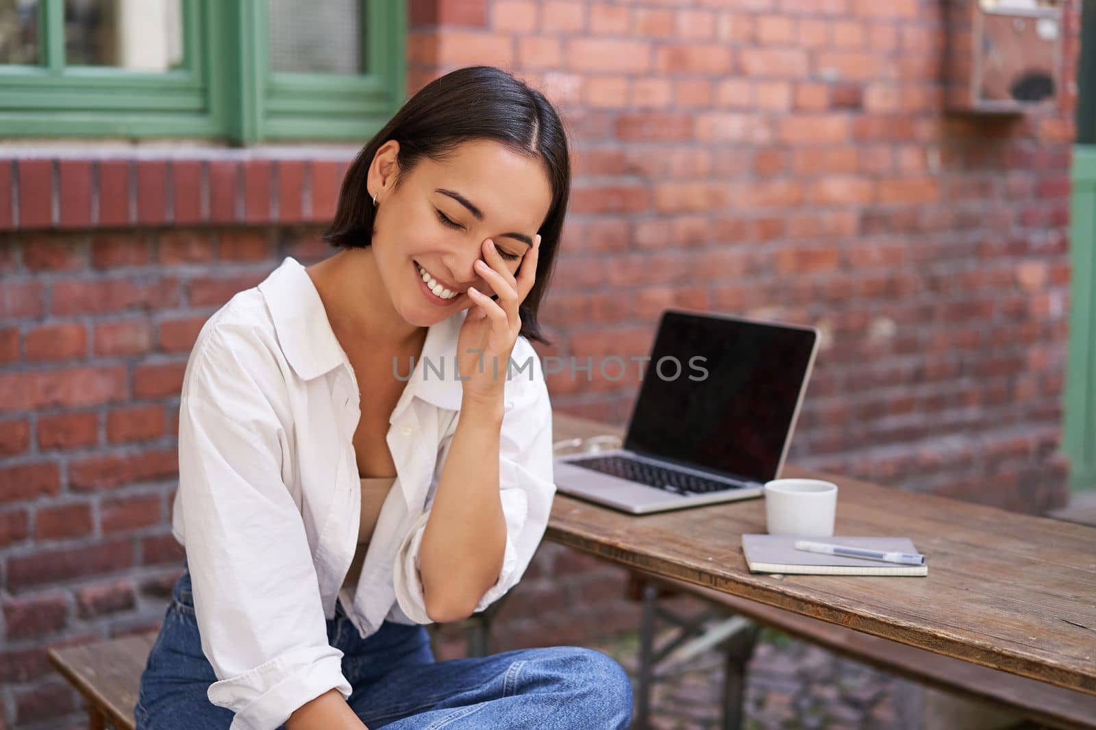 Beautiful korean girl with laptop, sitting in outdoor cafe, drinking coffee and using computer. Young people and recruitment concept