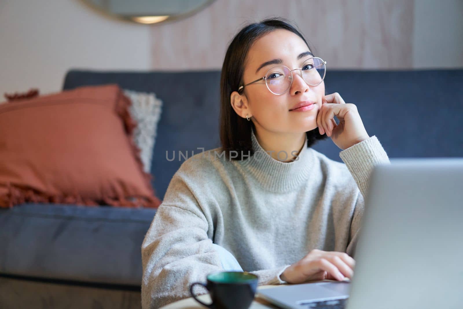 Self-employed young smiling woman, freelancer staying at home, working on remote from laptop, wearing glasses, sitting in living room.