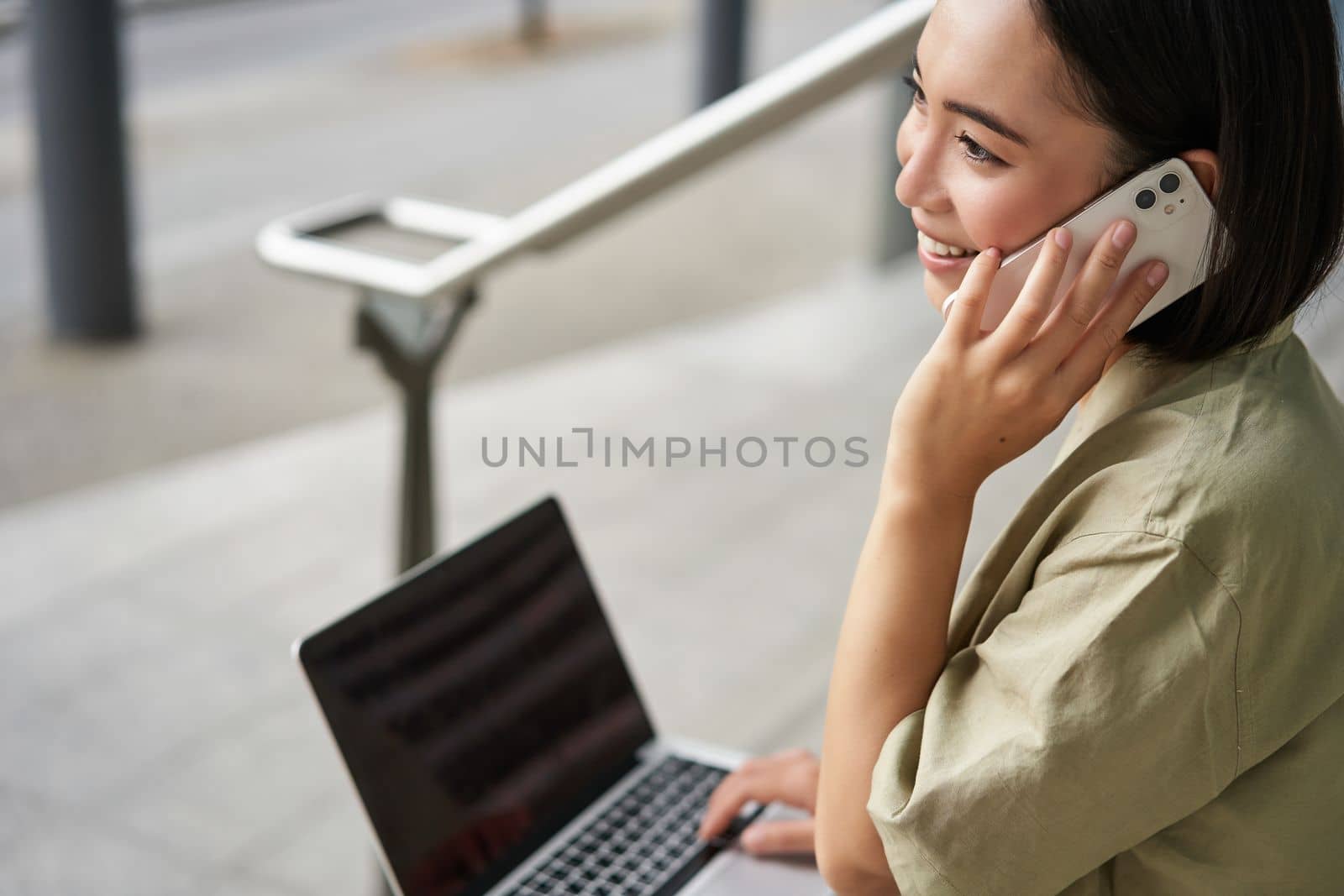 Portrait of asian girl, student sits on stairs with laptop, talks on mobile phone. Young woman makes a telephone call while working on computer.