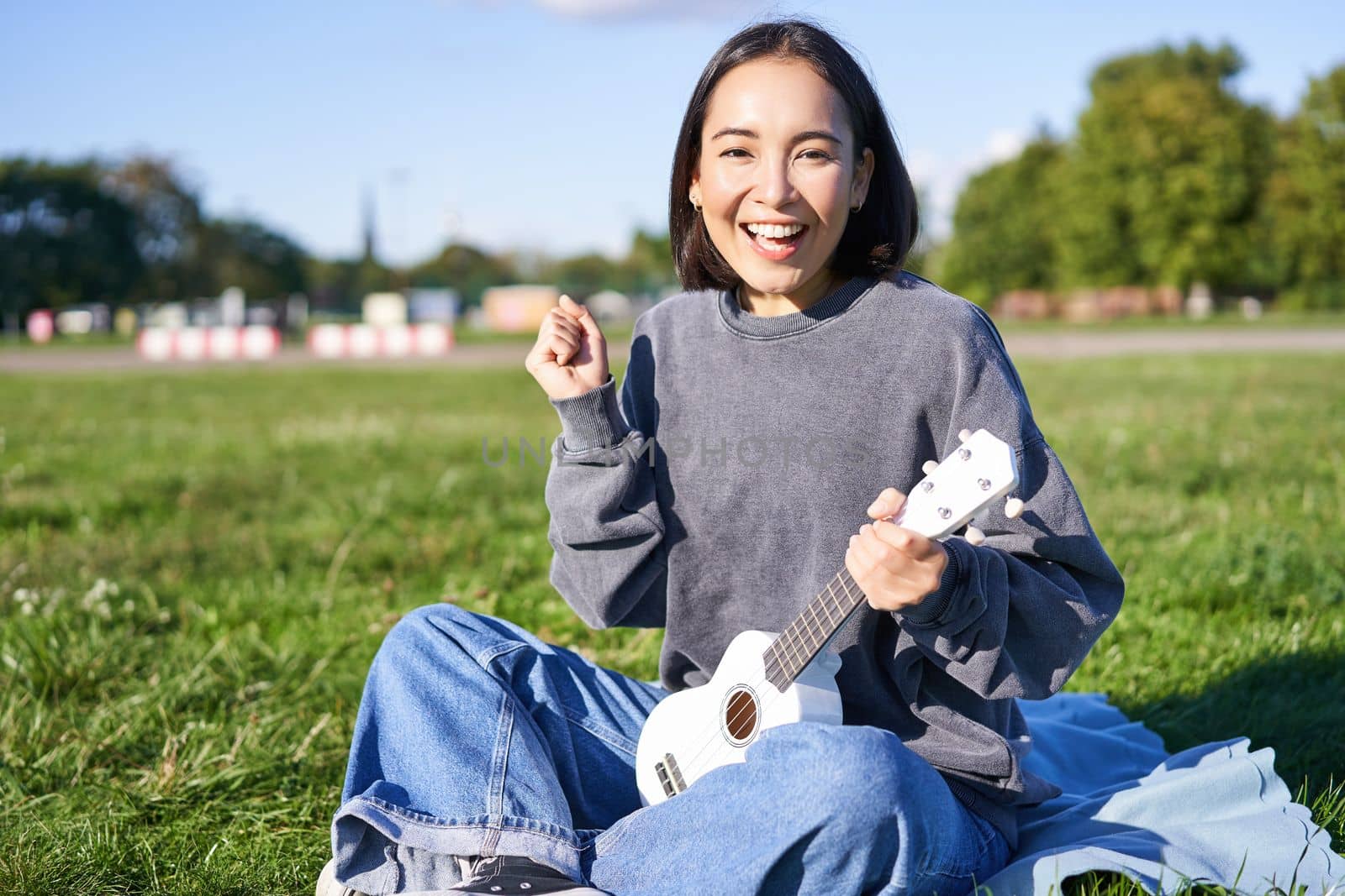 Portrait of excited asian girl, sitting with ukulele in park on green grass, looking amazed and surprised with good news.