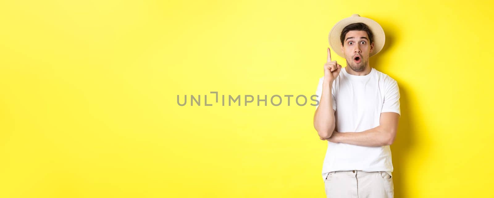 Portrait of young man in straw hat having an idea, raising finger eureka sign, making suggestion, standing over yellow background.