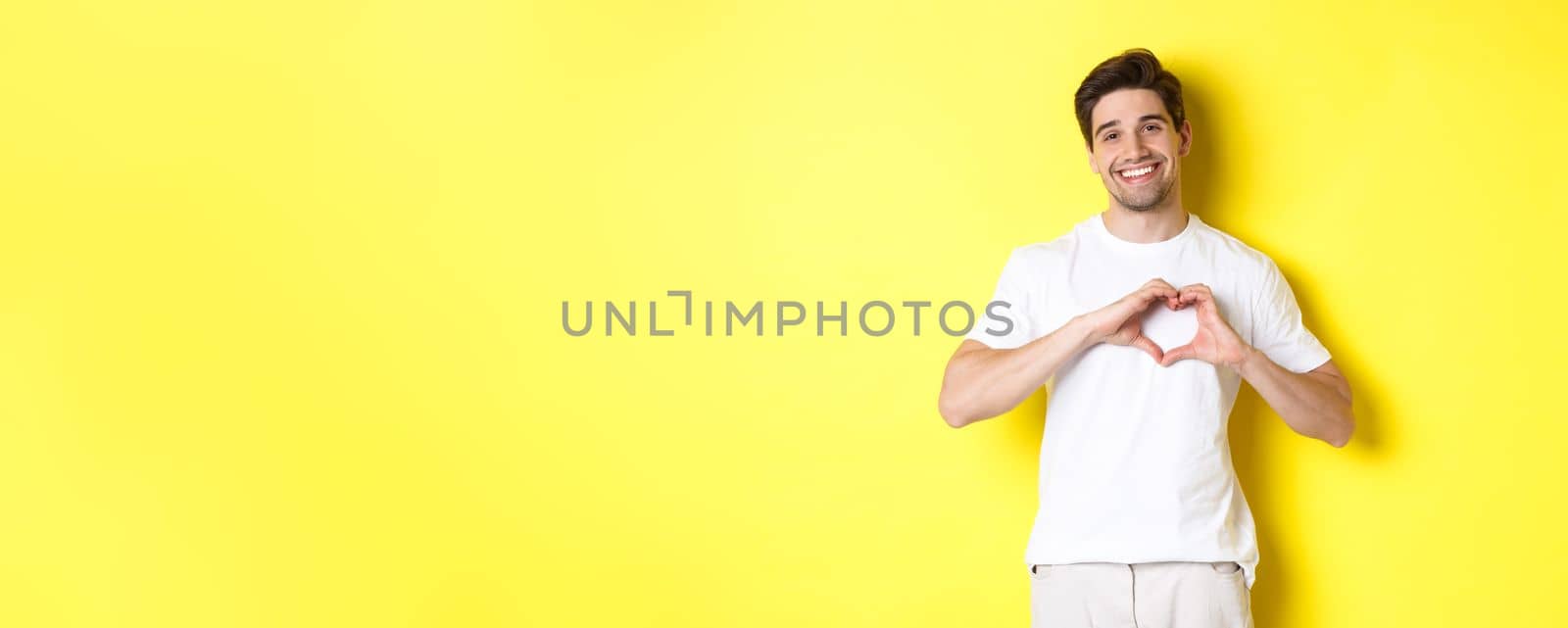 Happy romantic man showing heart sign, smiling and express love, standing over yellow background. Copy space