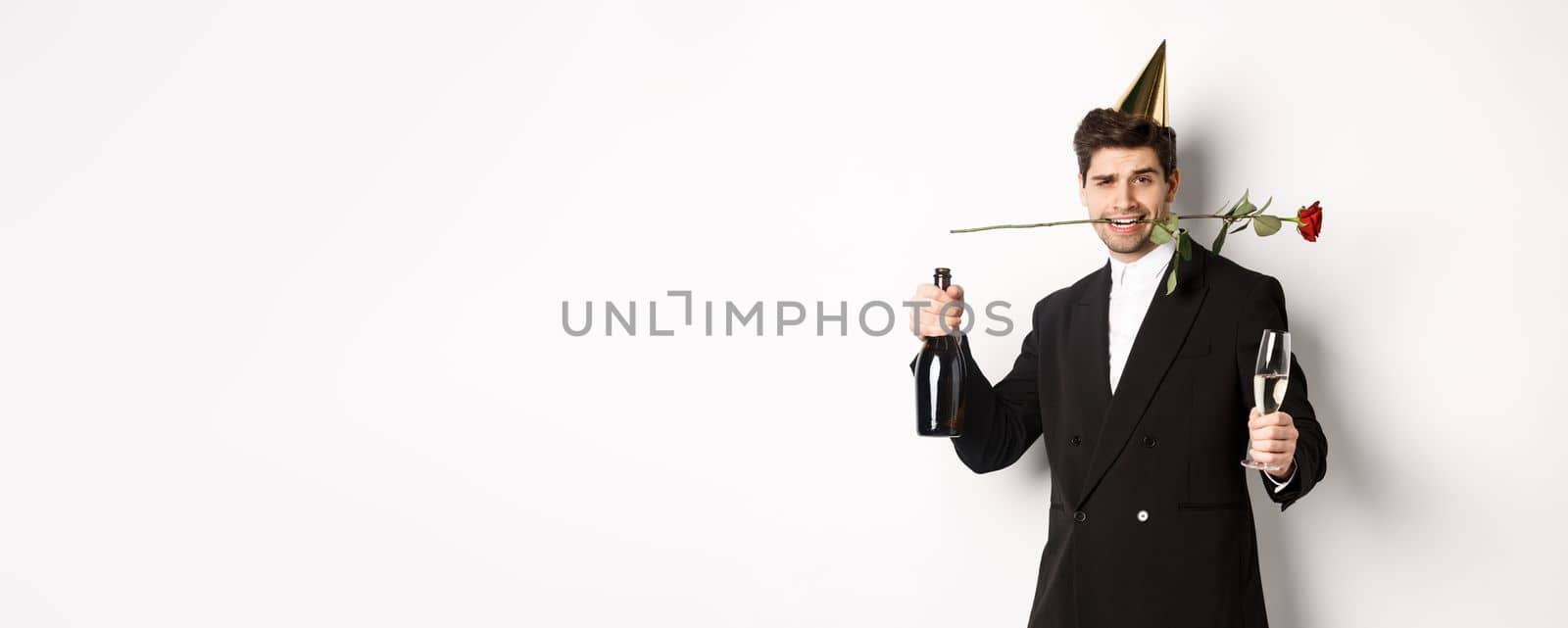 Romantic guy in trendy suit, celebrating and having a party, holding rose in teeth and champagne, standing over white background.
