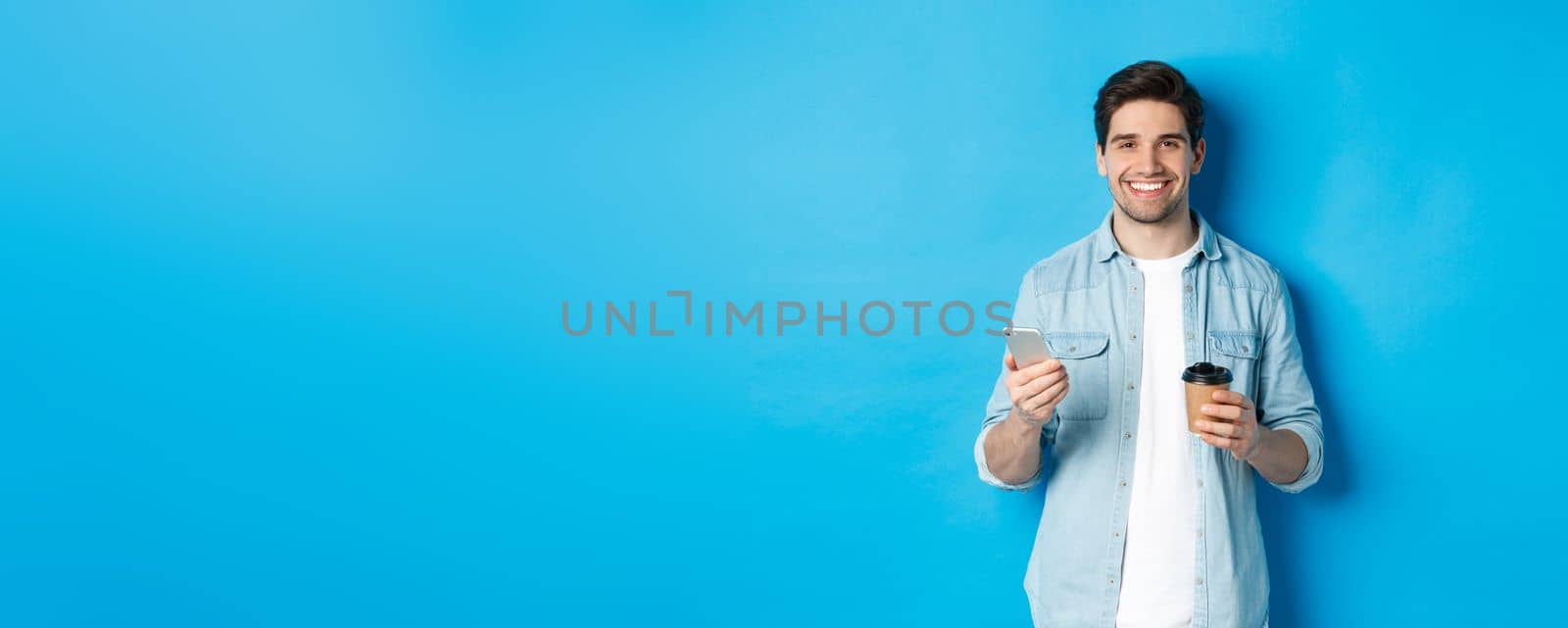 Young modern man drinking coffee and using mobile phone, smiling at camera, standing over blue background.