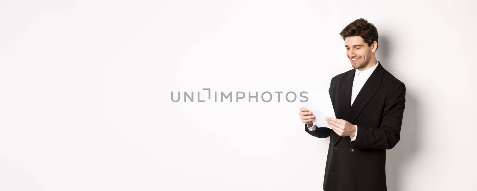 Image of attractive businessman in black suit, reading document and smiling, working on report, standing against white background.