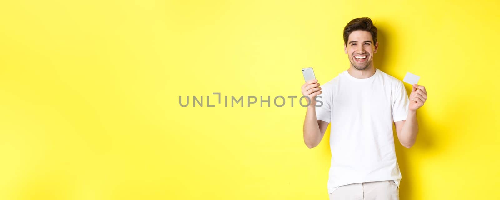 Happy young man shopping online in smartphone, holding credit card and smiling, standing over yellow background.