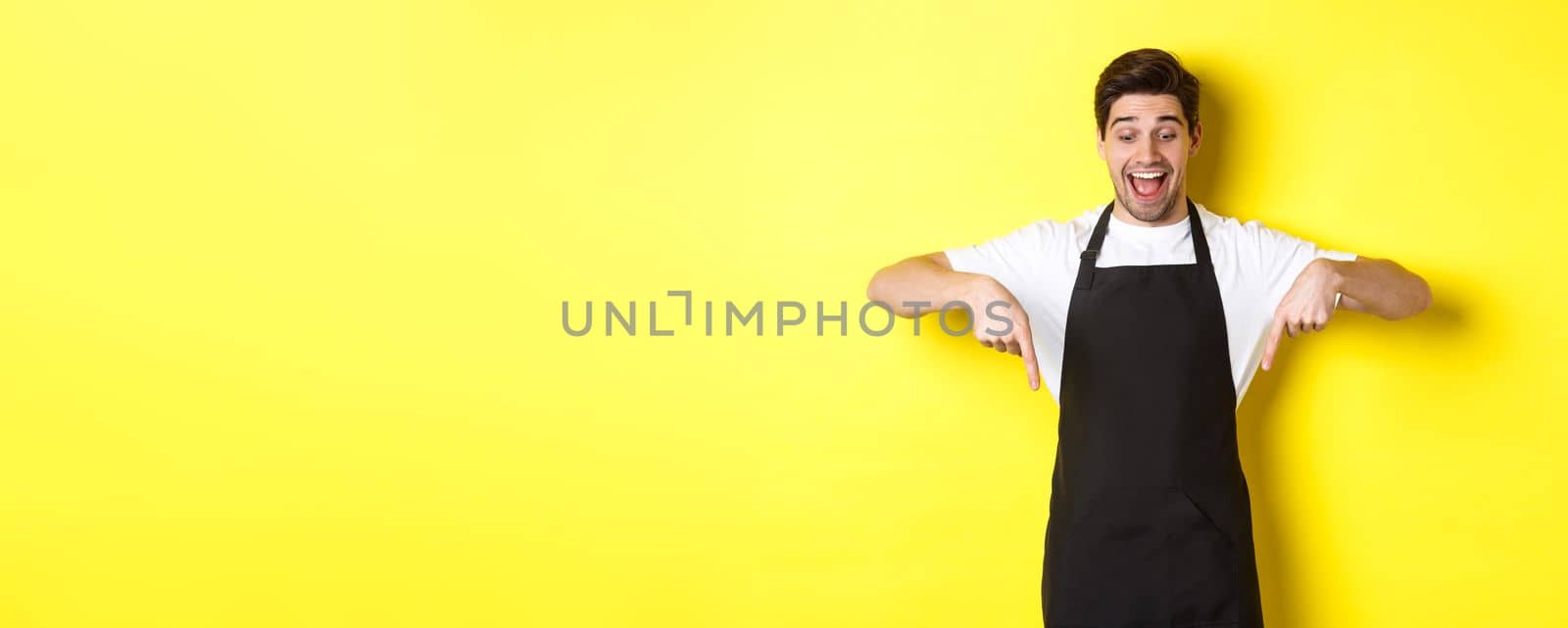 Excited waiter in black apron pointing fingers down, checking out promo offer, standing over yellow background.