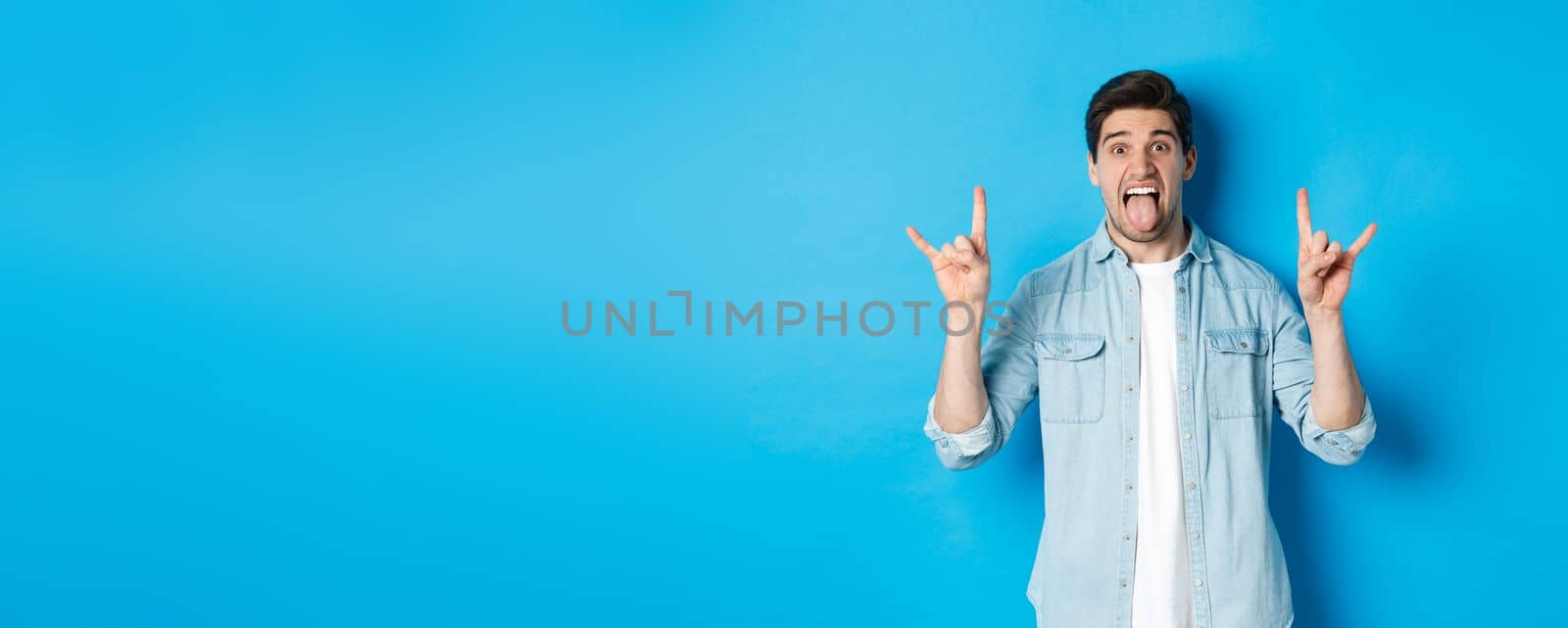 Cool guy having fun, enjoying concert, showing rock sign and tongue, standing over blue background.