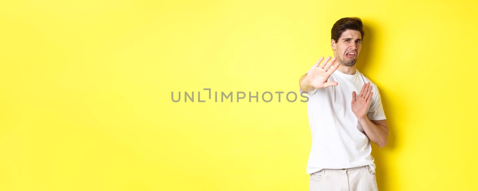 Disgusted man refusing, grimacing from dislike and aversion, begging to stop, standing in white t-shirt against yellow background by Benzoix