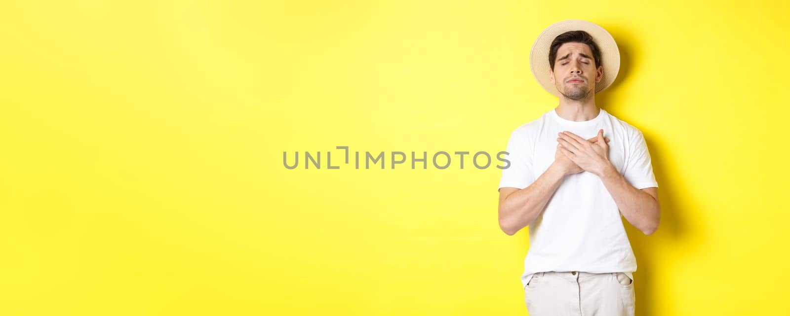 Concept of tourism and summer. Romantic man in straw hat looking nostalgic, close eyes and holding hands on heart, standing against yellow background.