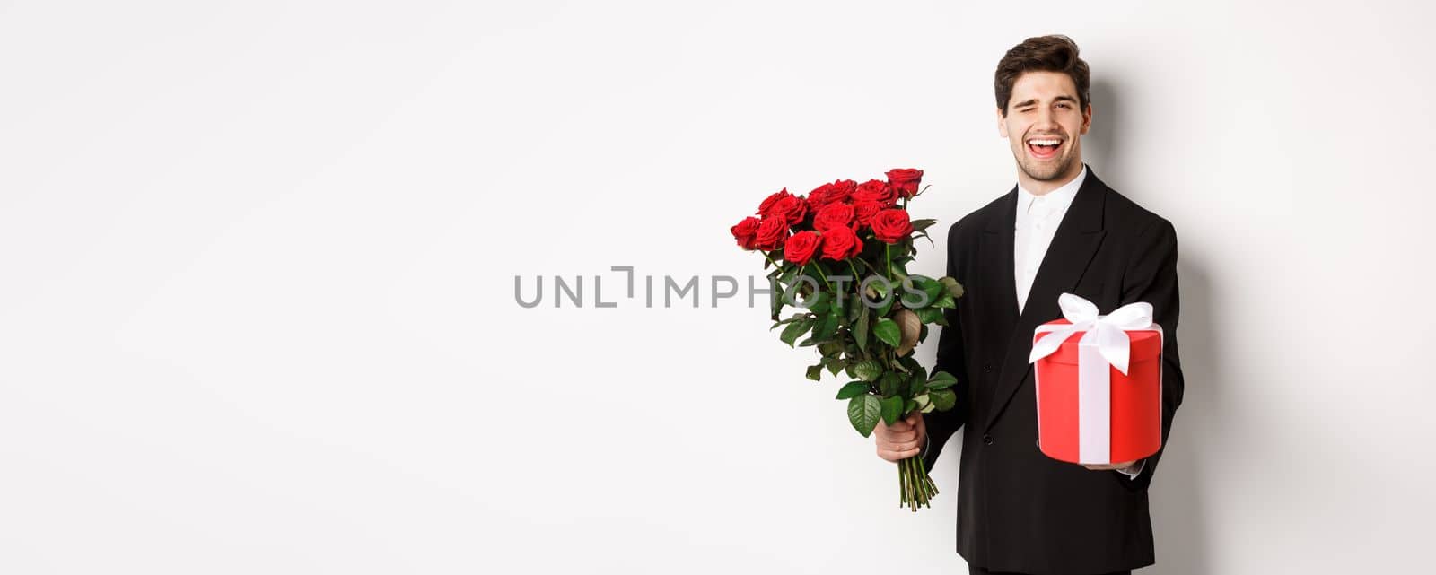 Concept of holidays, relationship and celebration. Charming young man in black suit, holding gift box and bouquet of roses, winking and smiling, standing against white background.