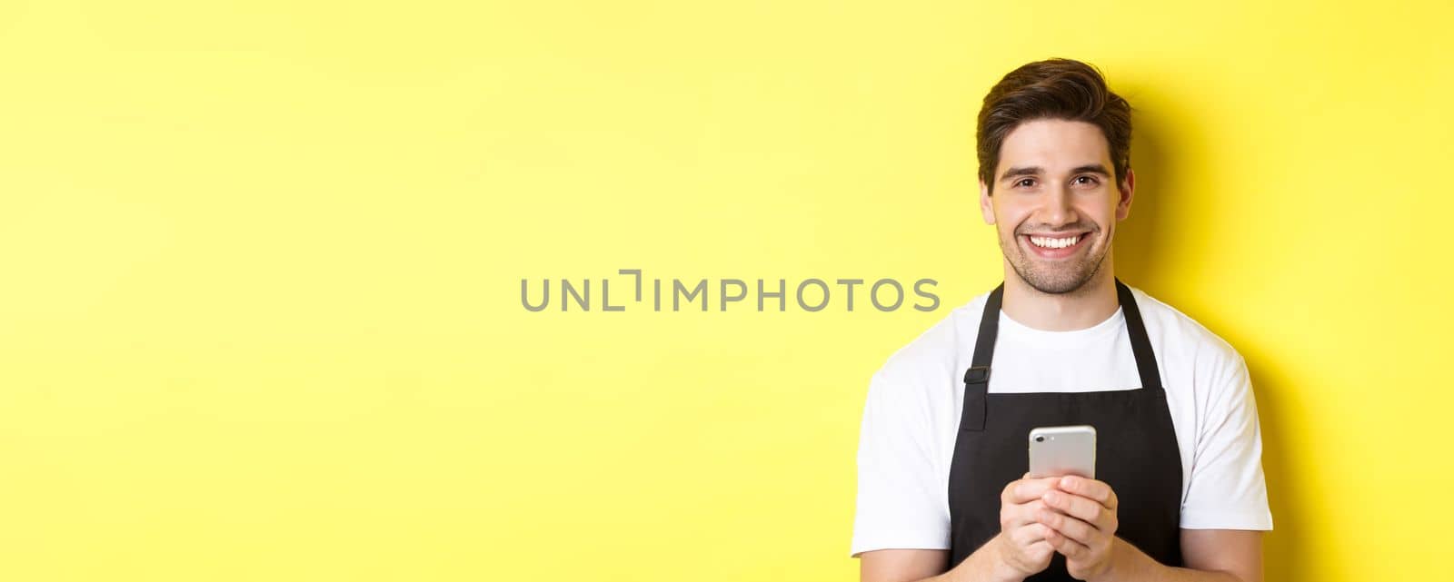 Close-up of handsome barista sending message on mobile phone, smiling happy, standing over yellow background.