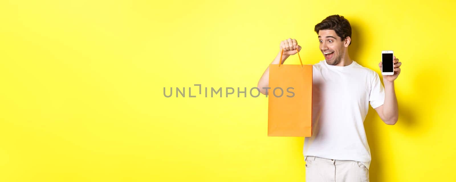 Concept of discounts, online banking and cashback. Happy guy buy something in store and looking at shopping bag, showing mobile phone screen, yellow background.