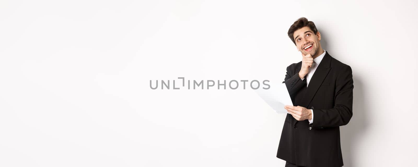 Image of thoughtful, handsome businessman in black suit, holding document and looking at upper right corner, thinking and smiling, standing against white background.