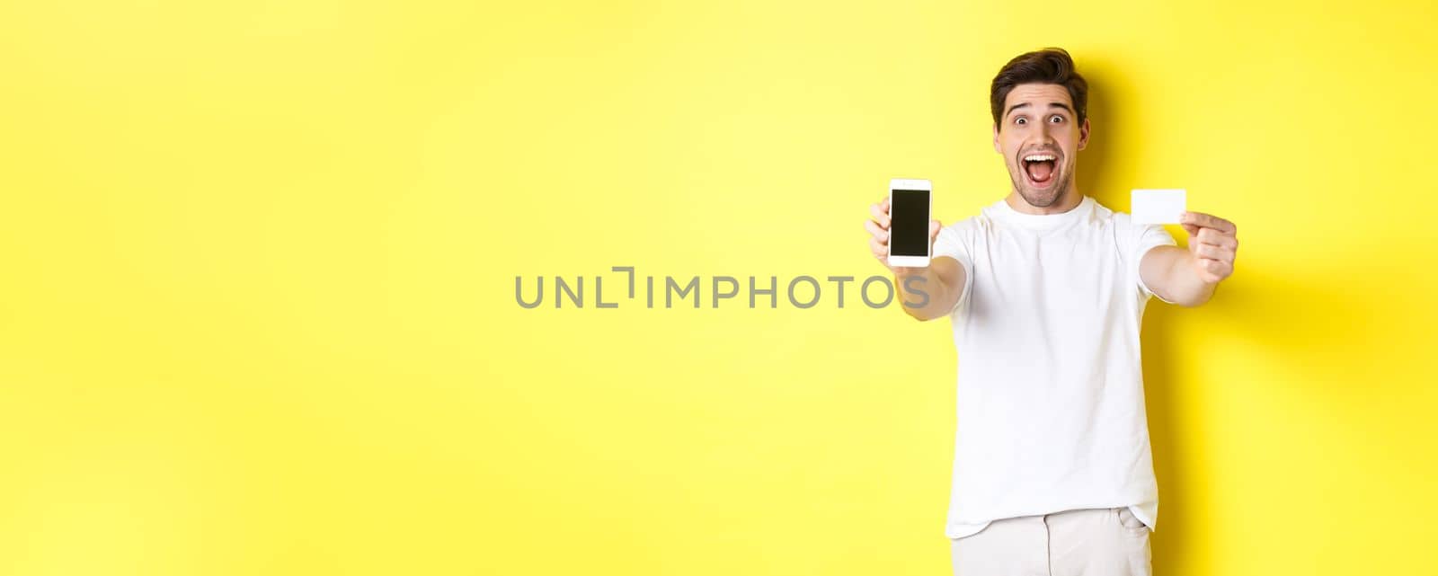 Handsome caucasian man showing smartphone screen and credit card, concept of mobile banking and online shopping, yellow background.