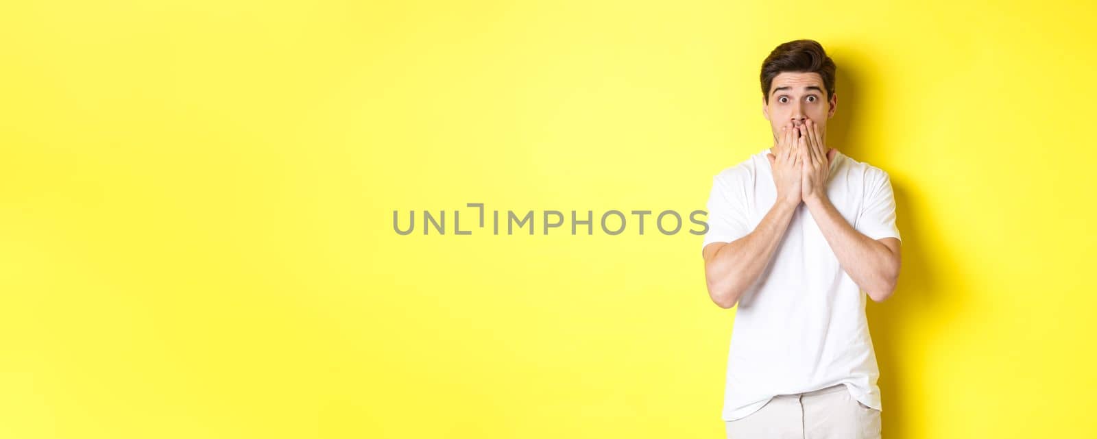 Handsome man looking shocked and speechless, holding hands on mouth, standing over yellow background by Benzoix