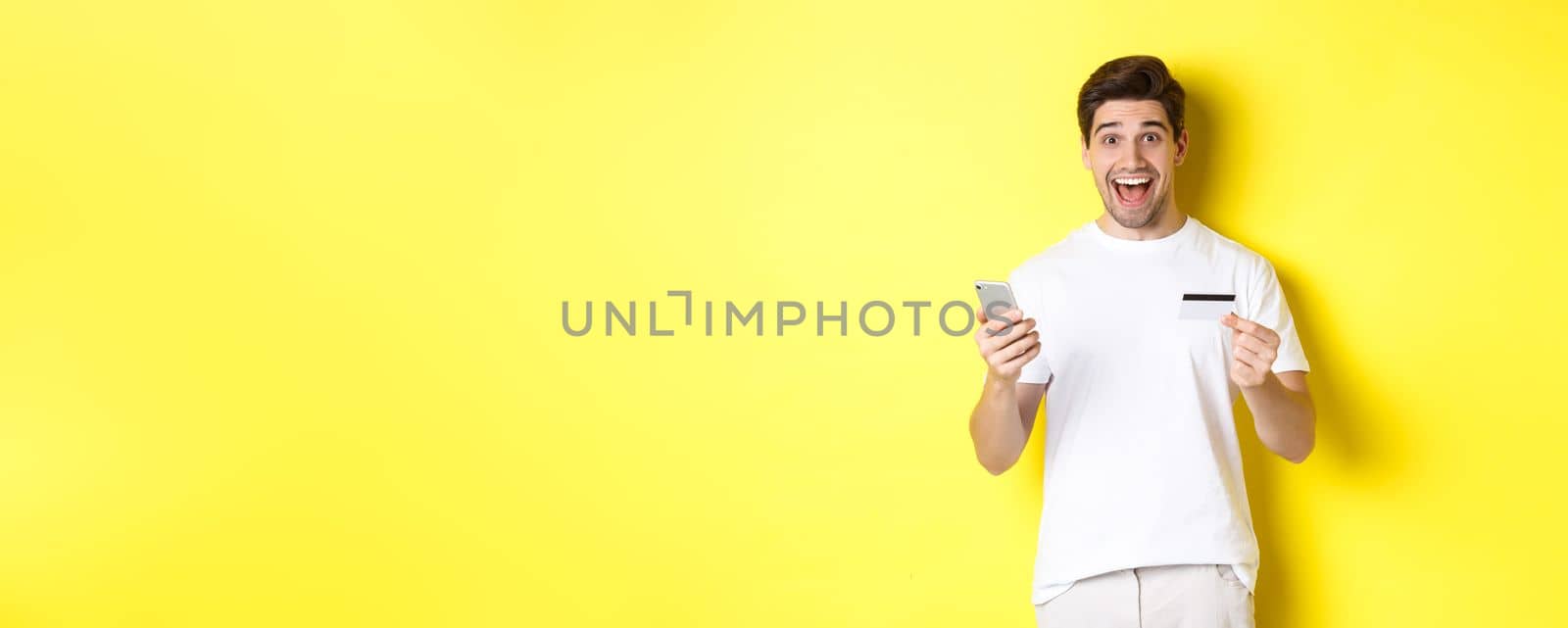 Happy male buyer holding smartphone and credit card, concept of online shopping in internet, standing over yellow background by Benzoix