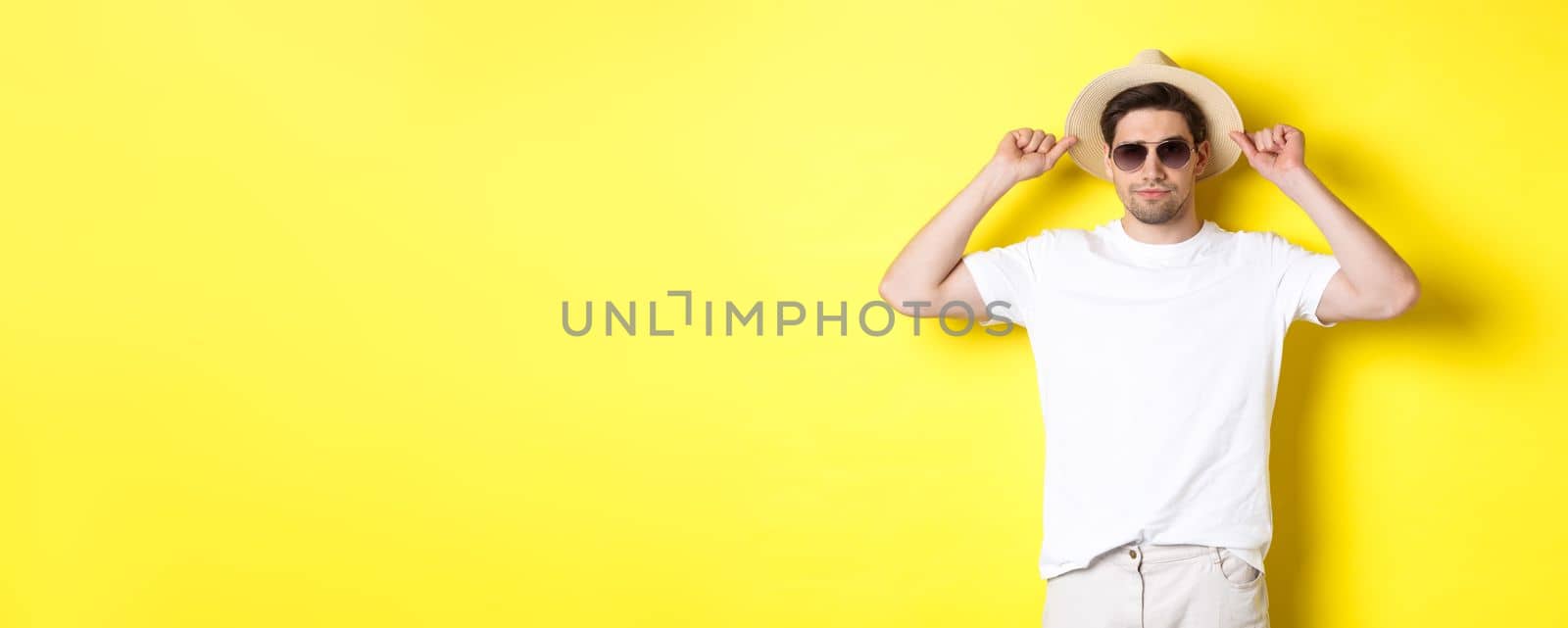Confident young male tourist ready for vacation, wearing straw hat and sunglasses, standing against yellow background by Benzoix