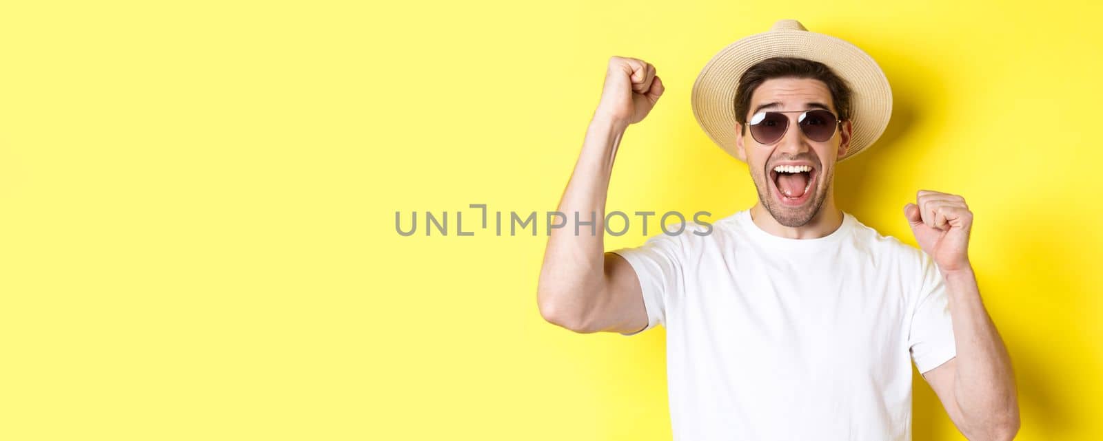 Concept of tourism and holidays. Happy male tourist celebrating his vacation, raising hands up and shouting for joy, wearing sunglasses with straw hat.