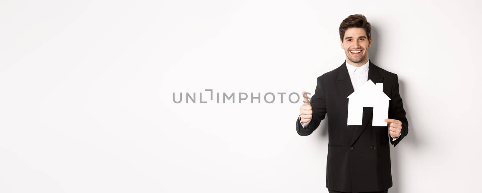 Portrait of confident real estate agent showing house maket and thumb-up, standing against white background.