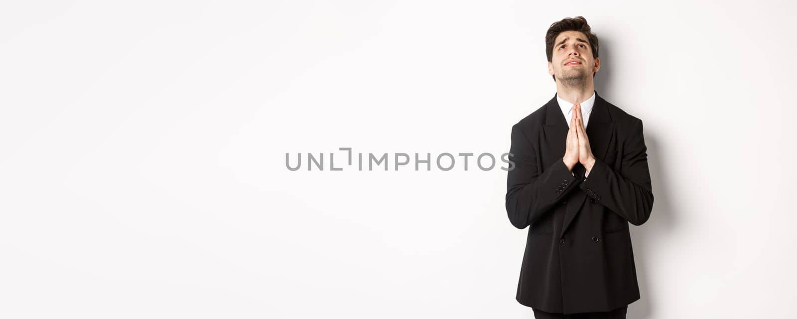 Troubled and hopeful man in black suit begging god, pleading and looking up, need help, standing over white background.