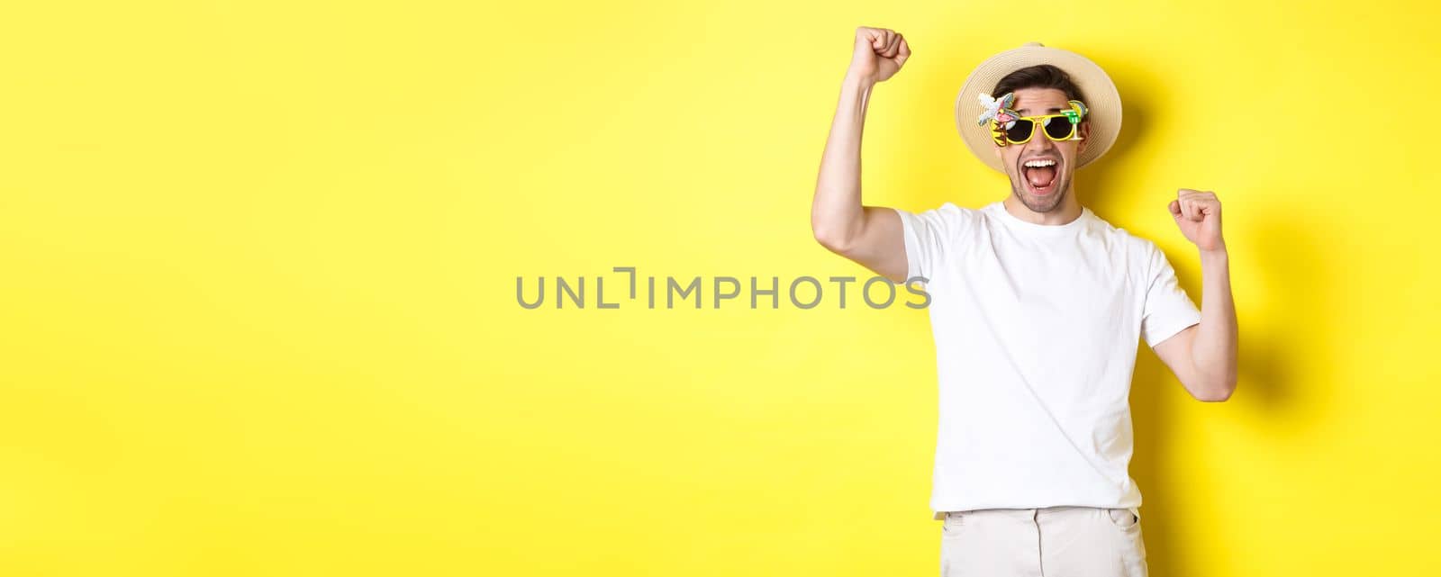Concept of tourism and lifestyle. Happy man winning trip to resort, shouting yes and raising hands up, triumphing, wearing sunglasses and summer hat, yellow background.