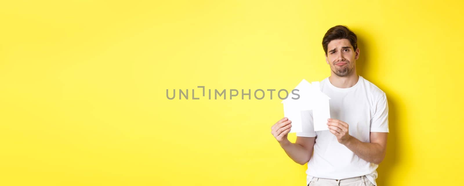 Real estate concept. Displeased young man showing paper house model and grimacing upset, standing over yellow background by Benzoix