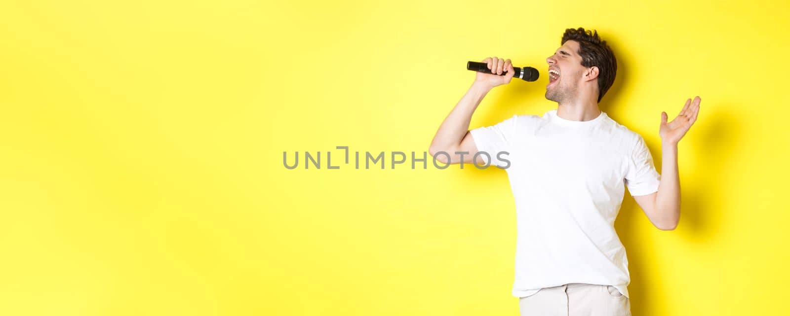 Young man singer holding microphone, reaching high note and singing karaoke, standing over yellow background by Benzoix
