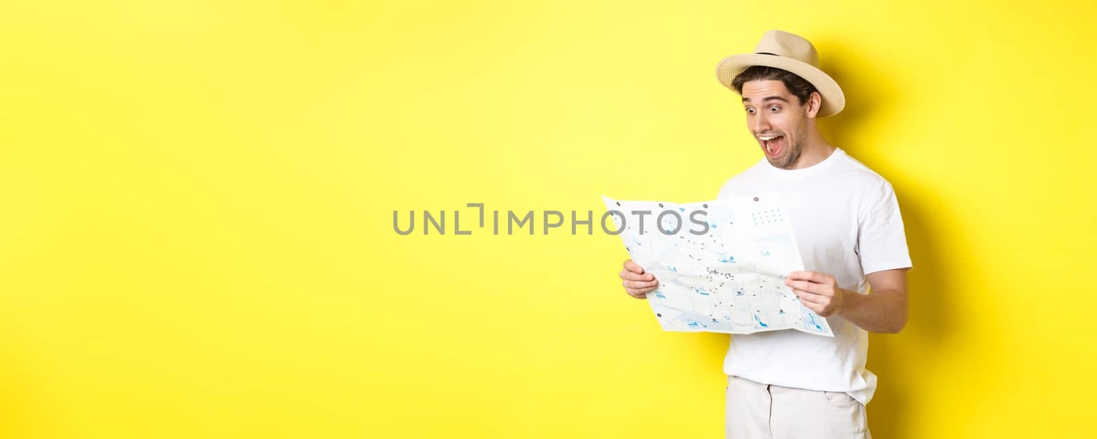 Travelling, vacation and tourism concept. Smiling happy tourist looking at map with sightseeings, standing against yellow background.