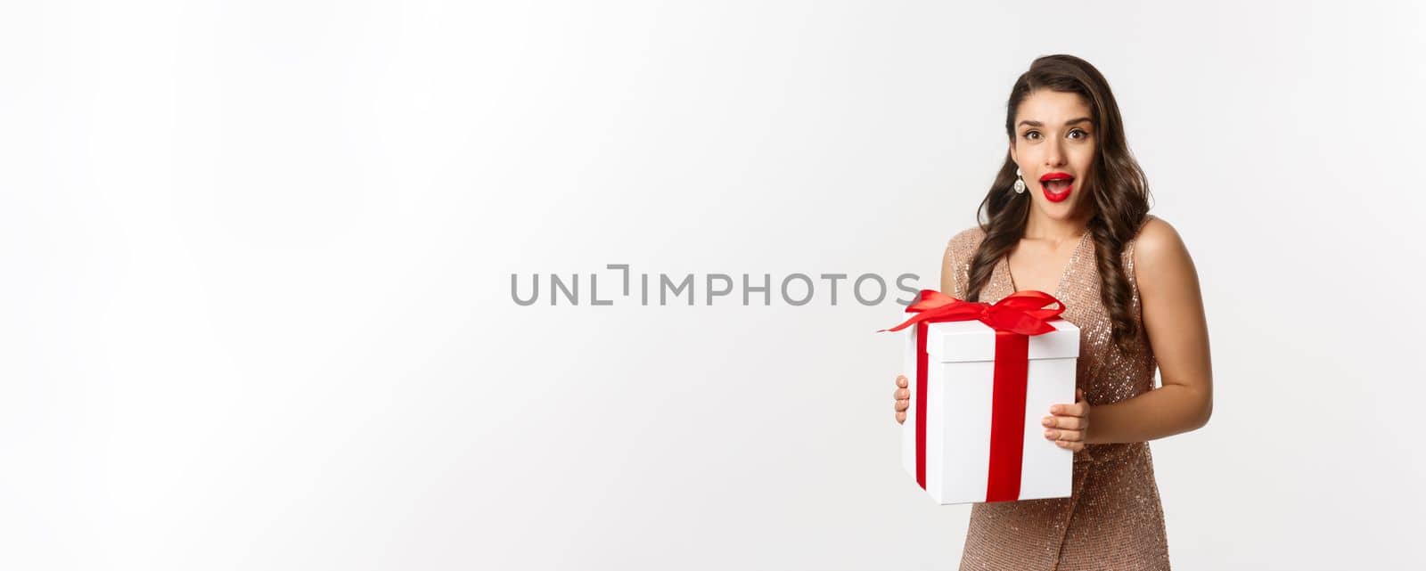 Merry Christmas. Image of beautiful woman in glamour dress receiving gift and looking surprised, celebrating New Year, standing over white background by Benzoix