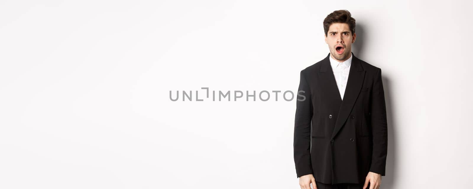 Portrait of shocked and startled handsome man in suit, drop jaw and looking in awe at camera, standing over white background.