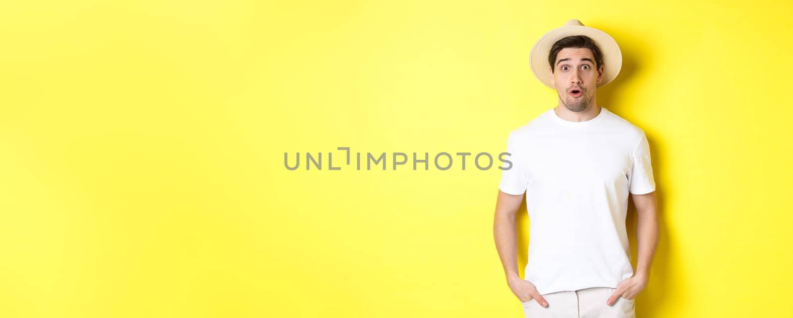 Concept of travelling and tourism. Surprised caucasian guy in straw hat, say wow, standing against yellow background by Benzoix