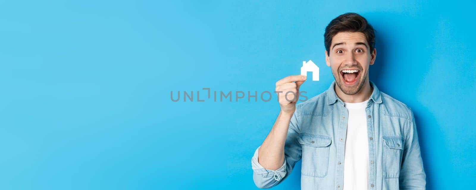 Real estate concept. Happy young man looking excited, found apartment, showing small house model, standing over blue background by Benzoix