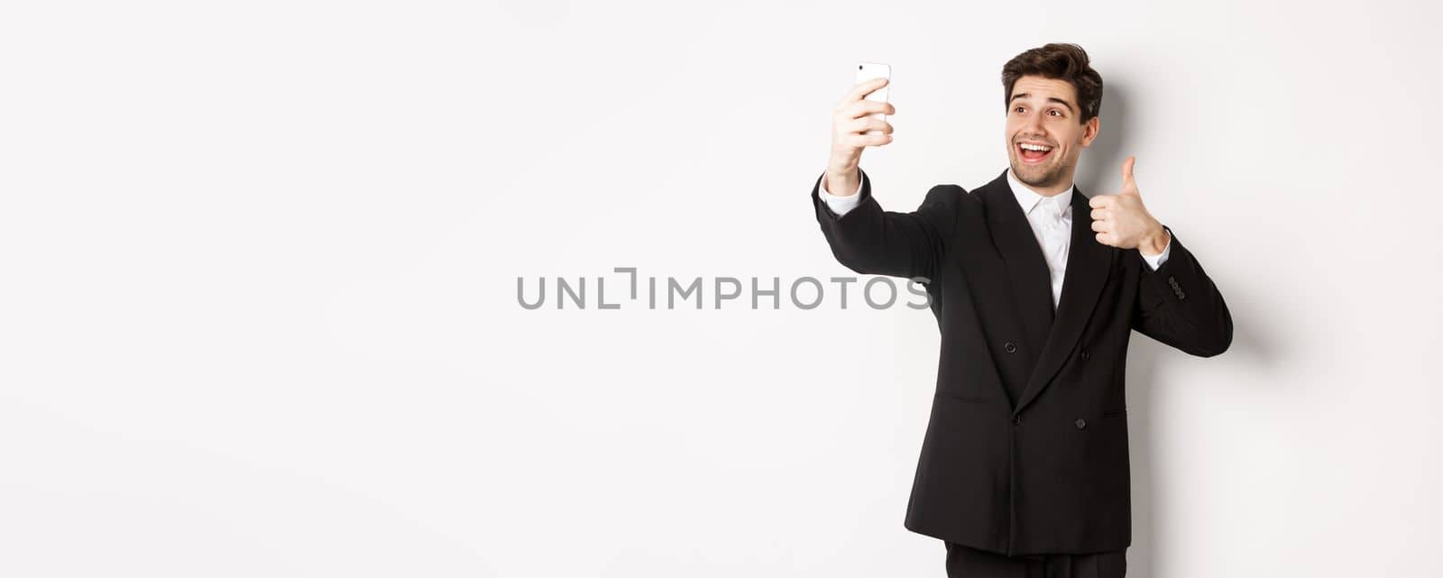 Portrait of good-looking man taking selfie on new year party, wearing suit, taking photo on smartphone and showing thumbs-up, standing against white background.