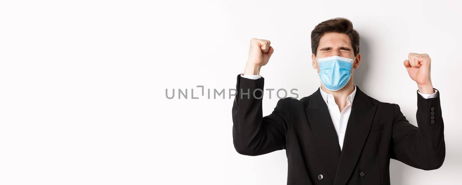 Concept of covid-19, business and social distancing. Close-up of handsome man in suit and medical mask, rejoicing and winning, raising hands up, shouting yes.