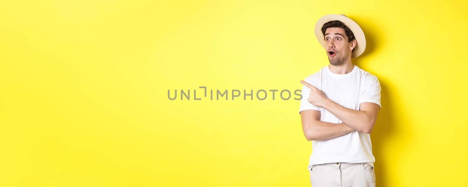 Concept of tourism and lifestyle. Excited handsome guy in straw hat checking out advertisement, pointing and looking at upper left corner logo, yellow background.