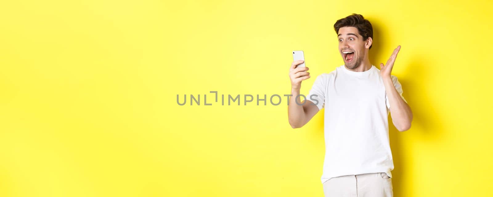 Surprised and happy man looking at mobile phone screen, reading fantastic news, standing over yellow background.