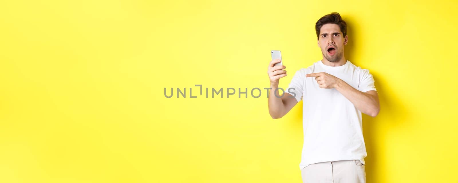 Confused man pointing finger at mobile phone screen, cant understand something, standing over yellow background by Benzoix