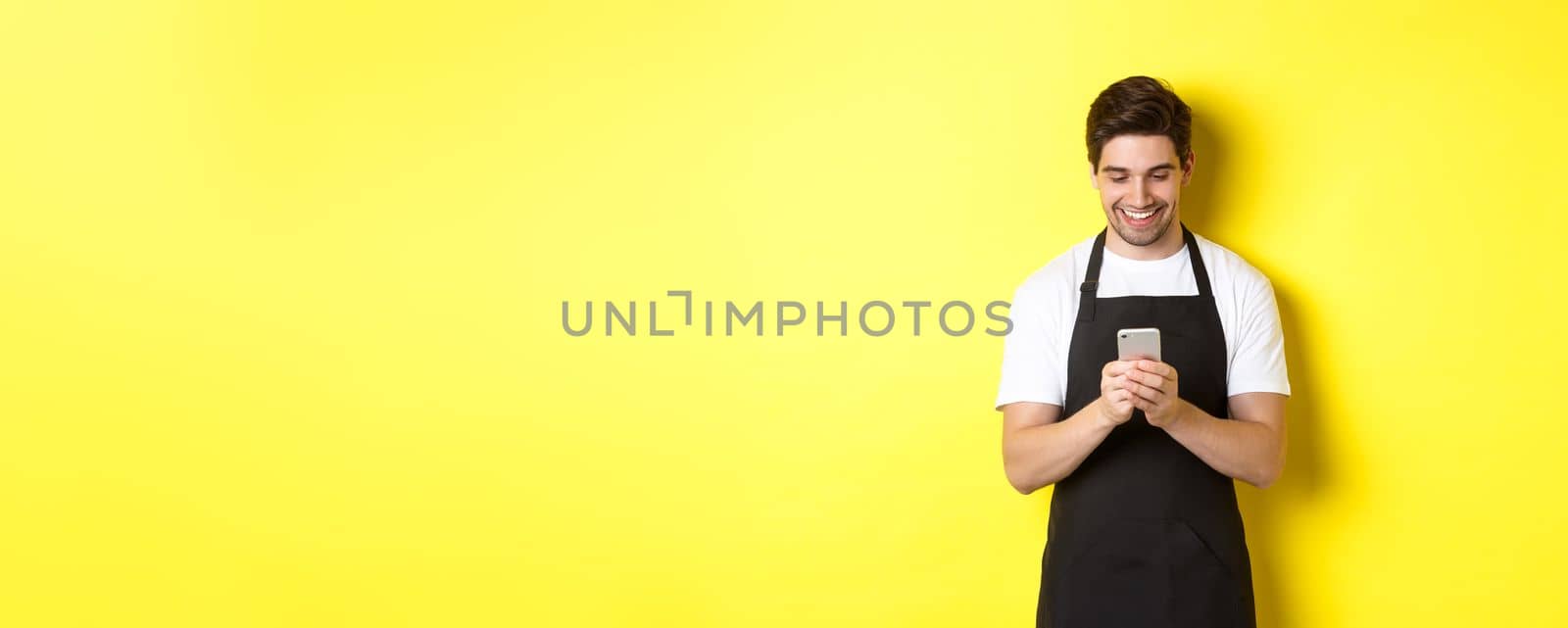 Waiter in black apron reading message on mobile phone, smiling happy, standing over yellow background by Benzoix