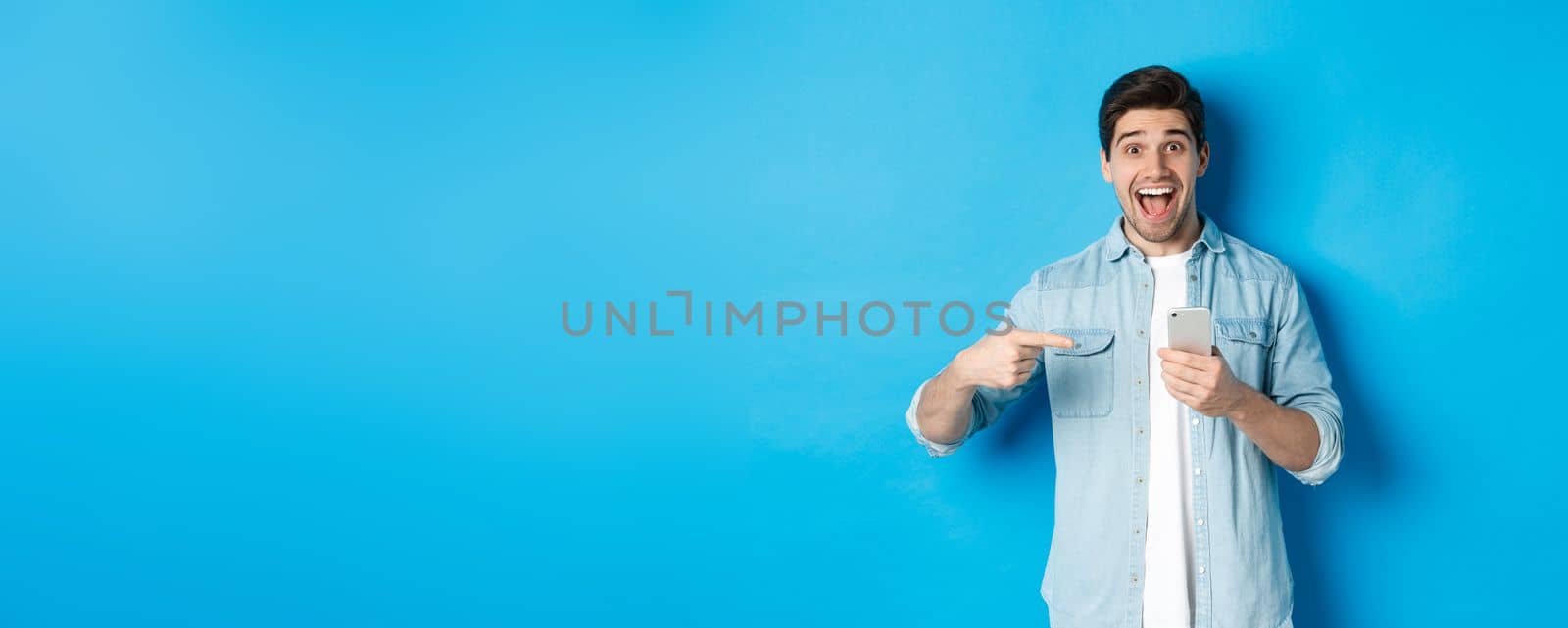 Concept of online shopping, applications and technology. Happy smiling man pointing finger at phone, standing amazed against blue background by Benzoix