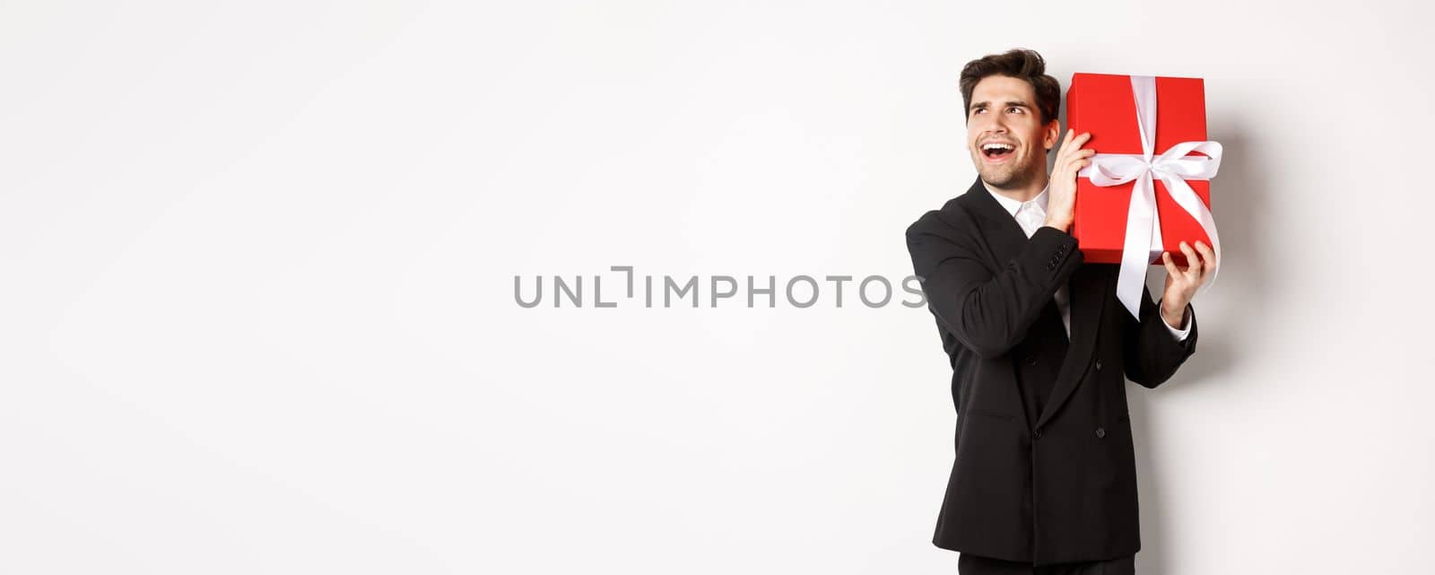 Concept of christmas holidays, celebration and lifestyle. Image of excited man enjoying new year, shaking gift box to guess what inside, standing against white background.