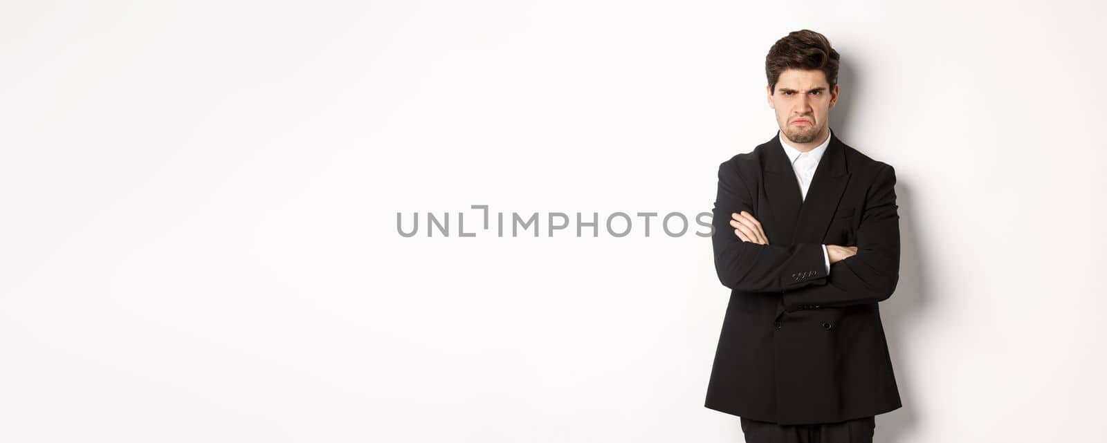 Portrait of angry handsome man in black suit, cross arms on chest and looking offended, frowning and pouting, being mad at someone, standing over white background.