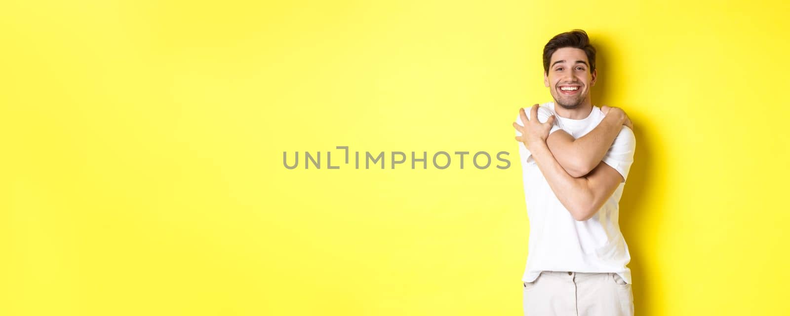 Happy kind guy hugging himself and smiling, standing pleased against yellow background. Copy space